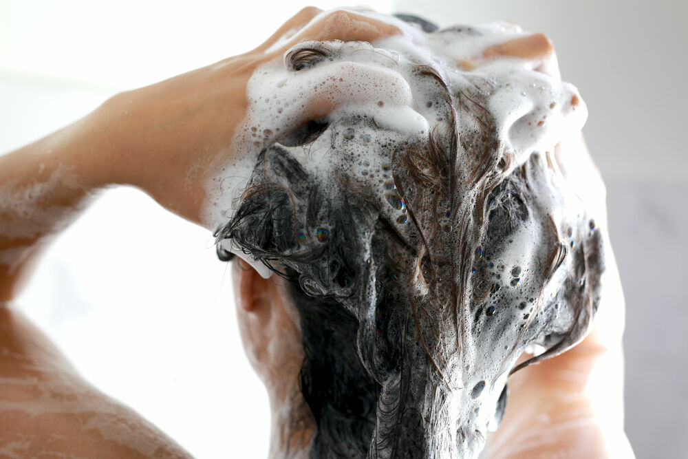 Osoba myje głowę, masując włosy pokryte obfitą pianą z szamponu. Widać tylko ręce i umyte włosy.