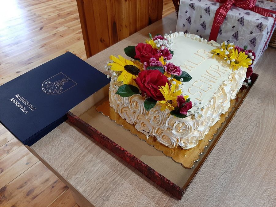 
                                                    Zdjęcie przedstawia tort
udekorowany żywymi kwiatami w kolorze żółtym, czerwonym i różowym
z napisem Klub Senior+
                                                