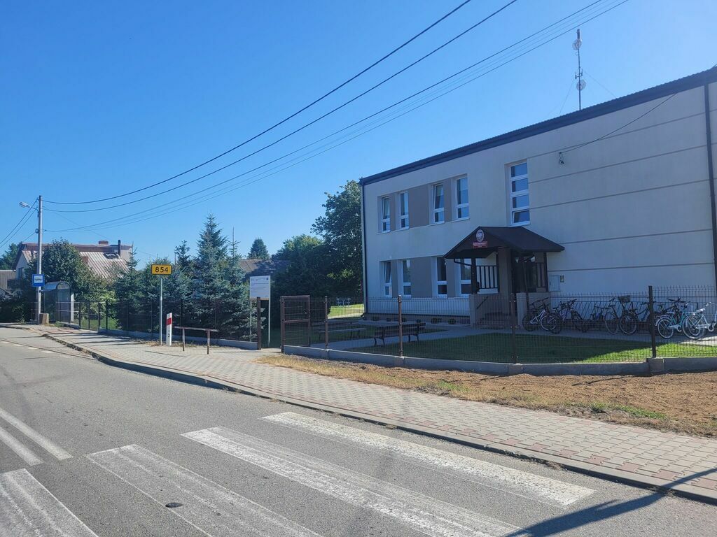 
                                                    Zdjęcie przedstawia jednopiętrowy budynek szkolny z ciemnoszarym dachem, otoczony niewysokim metalowym ogrodzeniem, przy słonecznym dniu. Obok szkoły widoczny rowerowy stojak z rowerami oraz drogowskaz przy drodze.
                                                