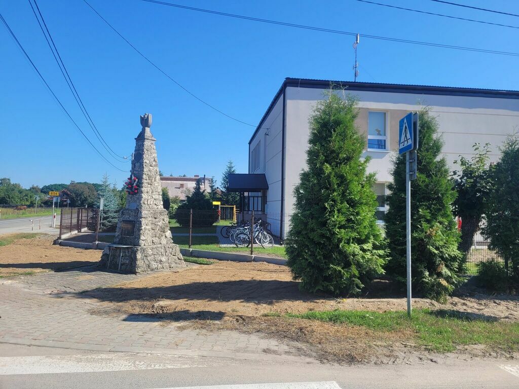 
                                                    Przed szkołą stoi kamienne ogrodzenie w formie niewielkiej wieży, obok rosną zielone tuje, po prawej widać rowery, w tle budynek szkolny i niebieskie niebo.
                                                