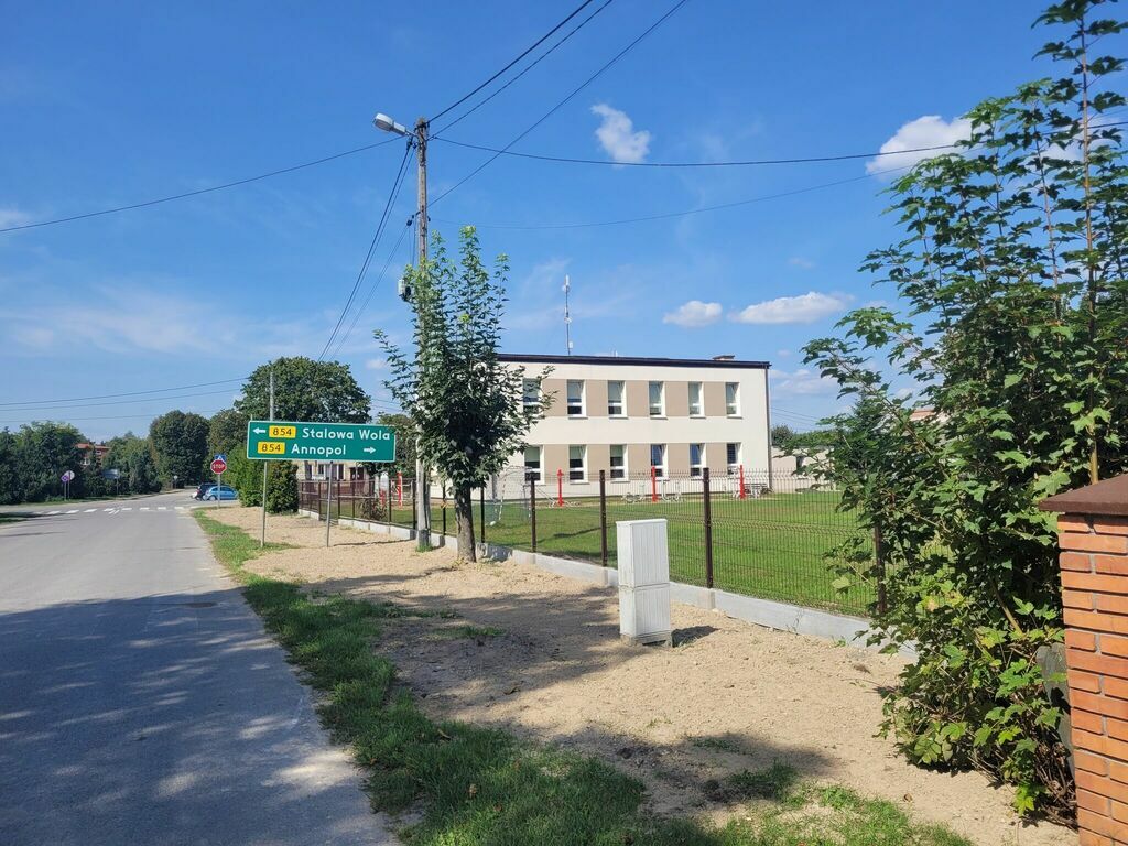 
                                                    Zdjęcie przedstawia szarą budowę szkoły z białymi oknami, za metalowym ogrodzeniem, z otoczeniem zieleni i niebieskim niebem, przy asfaltowej drodze z drogowskazem.
                                                