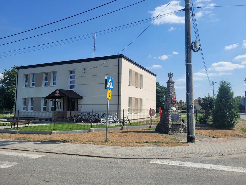 
                                                    Zdjęcie przedstawia budynek szkolny przy słonecznej pogodzie. Obok budynku widoczne ogrodzenie i pomnik, a także rowery zaparkowane przy wiacie. Na pierwszym planie droga i znak przejścia dla pieszych.
                                                