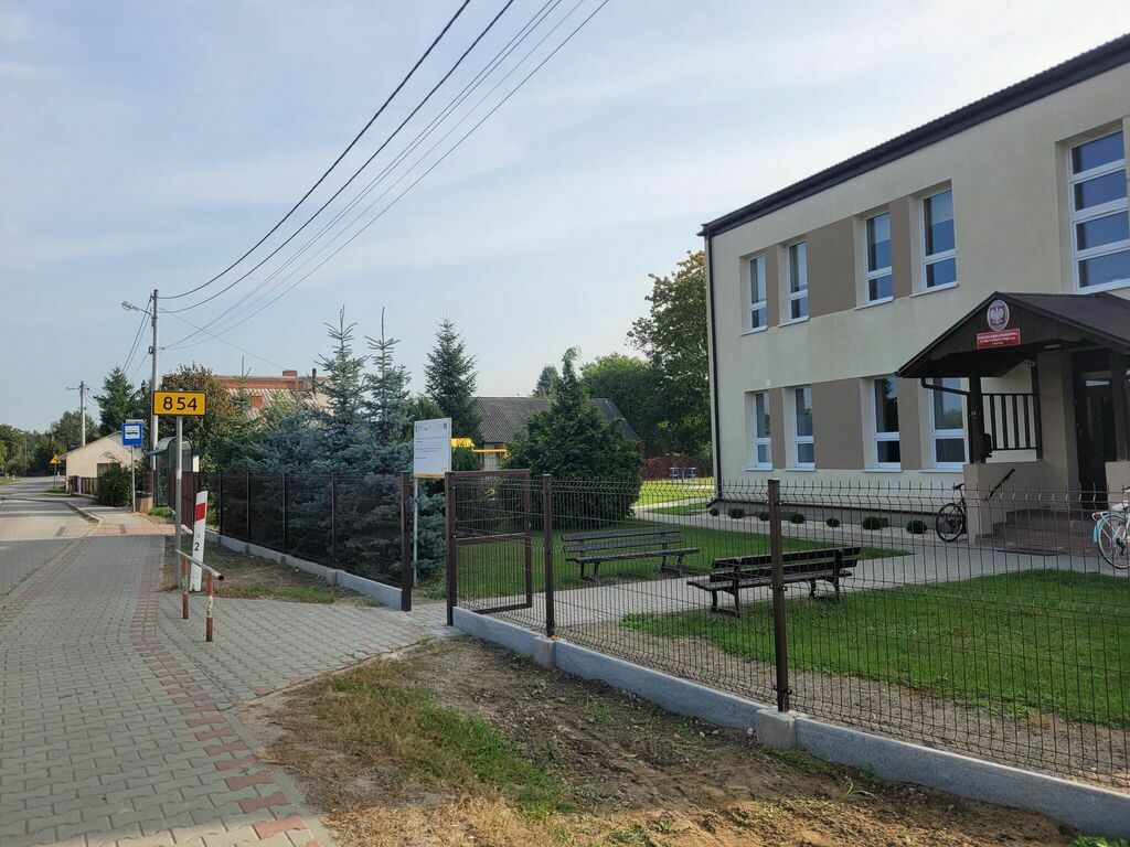 
                                                    Zdjęcie przedstawia szkołę z jasną fasadą widoczną po prawej stronie z przyległym ogrodzeniem i zielenią. Po lewej stronie jest droga i znak drogowy.
                                                