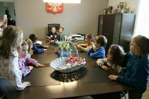 Dzieci siedzą przy długim biurku, na środku koszyk z lizakami, w tle półki z trofeami.