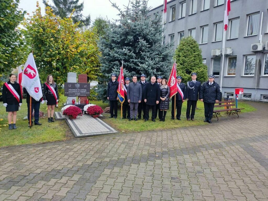 Grupa osób stoi przed kamieniem pamiątkowym z flagami Polski i sztandarami. Jest pochmurno. Obecni są mundurowi i cywile, oddając hołd.