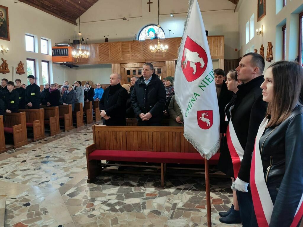 Wnętrze kościoła podczas uroczystości. Grupa osób stoi w rzędzie, niektóre w mundurach. Po prawej stronie flaga z herbem gminy.
