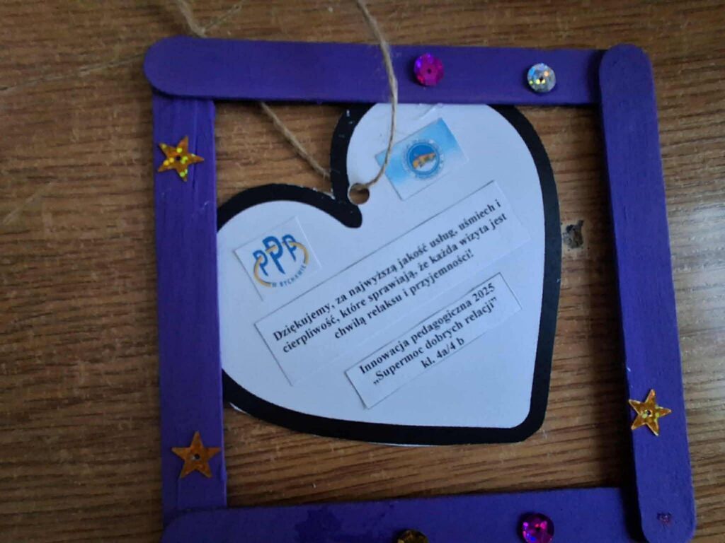 
                                                    Fioletowa ramka ozdobiona gwiazdkami i dżetami. W środku przypięto wycięte serduszko z tekstem podziękowania, logotypami oraz napisem „Innowacyjna placówka”. Leży na drewnianym tle.
                                                