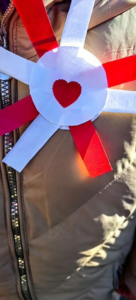 
                                                    Na zdjęciu widoczna jest papierowa odznaka w kształcie gwiazdy, z czerwonym sercem w środku. Odznaka jest przypięta do beżowej kurtki z błyszczącym zamkiem. Odznaka składa się z czerwonych i białych pasków papieru.
                                                