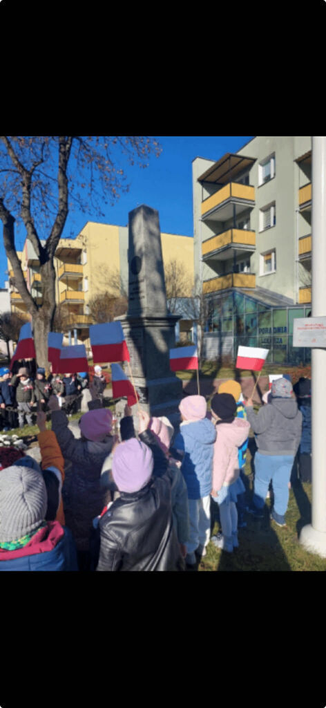 
                                                    Grupa dzieci trzymających polskie flagi stoi wokół pomnika na tle budynków. Dzieci są ciepło ubrane, co sugeruje chłodniejszą porę roku. W tle widać drzewa i elementy miejskiego krajobrazu.
                                                