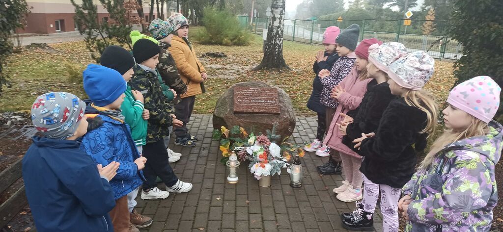 
                                                    Grupa dzieci stoi w kręgu na zewnątrz, patrząc na kamień pamiątkowy otoczony świecami i kwiatami. Dzieci są ciepło ubrane, a w tle widoczne są drzewa i ścieżka.
                                                