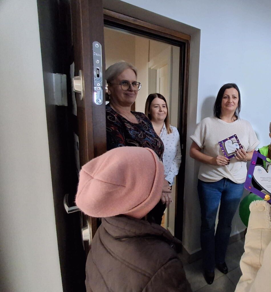 
                                                    Na zdjęciu wewnątrz budynku stoją trzy uśmiechnięte osoby, trzymające kolorowe teczki. Po lewej stronie w drzwiach znajduje się osoba w różowej czapce i kurtce, zwrócona tyłem do aparatu.
                                                