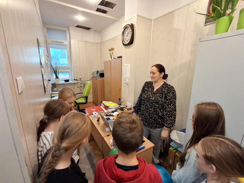 
                                                    Grupa dzieci stoi przy biurku, rozmawiając z dorosłą osobą. Na biurku znajdują się różne przedmioty biurowe. Ściany pomieszczenia są jasne, w tle widać krzesło, szafkę oraz okno.
                                                