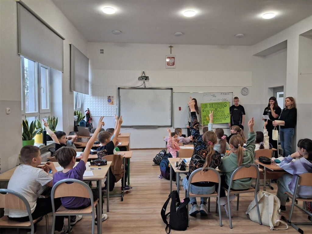 
                                                    Na zdjęciu widać grupę dzieci siedzących w klasie, większość z uniesionymi rękami. Na przodzie stoją dwie osoby przy tablicy, jedna trzyma kartkę z napisem. W tle widać tablicę i okna z roślinami na parapecie.
                                                