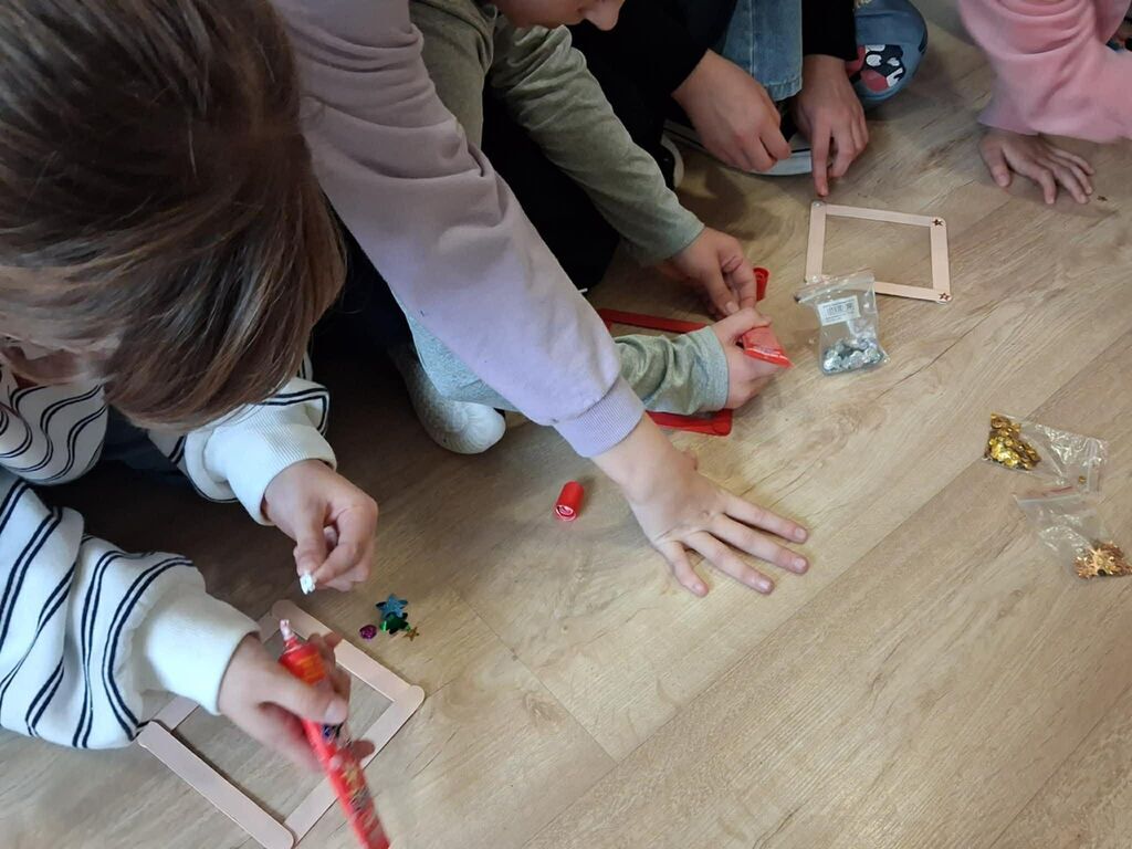 
                                                    Grupa dzieci siedzi na podłodze, dekorując ramki na zdjęcia. Używają kolorowych ozdób i kleju. Każde dziecko pracuje nad swoją ramką, skupiając się na szczegółach.
                                                