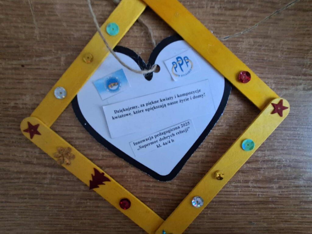 
                                                    Na zdjęciu widoczna jest dekoracyjna ramka z żółtymi patyczkami, ozdobiona kolorowymi naklejkami. Wewnątrz znajduje się karta z informacją i logo. Ramka wisi na sznurku na drewnianym tle.
                                                