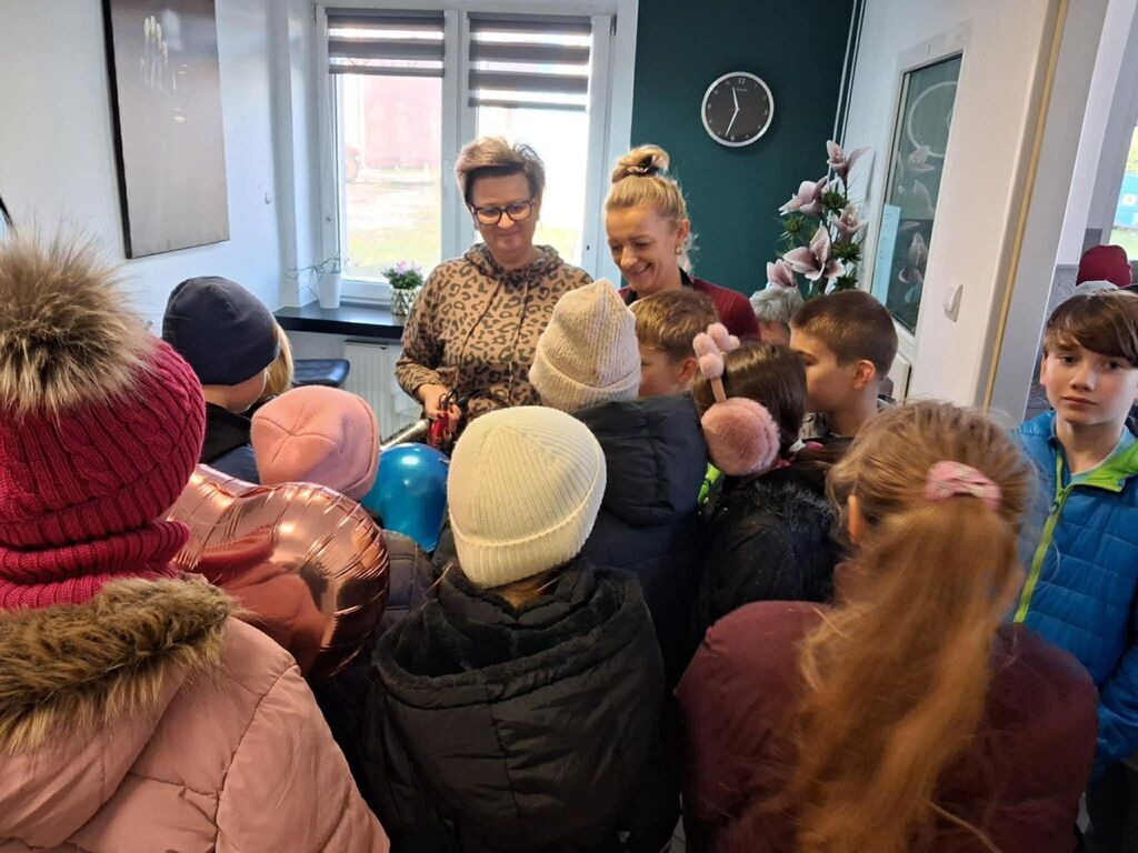 
                                                    Grupa dzieci i dwie osoby dorosłe stoją w pomieszczeniu. Dzieci są ubrane w ciepłe zimowe ubrania i patrzą na przedmiot trzymany przez jedną z dorosłych. W tle widać zegar i kwiaty.
                                                