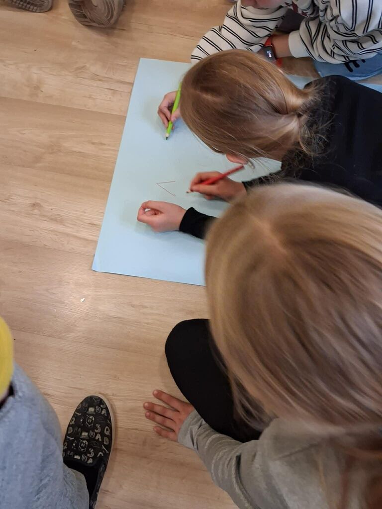 
                                                    Na zdjęciu widoczne są dwie osoby siedzące na podłodze. Jedna z nich pisze coś na dużym arkuszu niebieskiego papieru za pomocą czerwonego flamastera, a druga trzyma zielony flamaster. Obie są ubrane swobodnie.
                                                