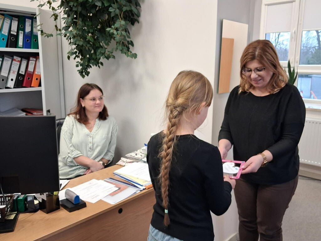 
                                                    Nie wiem, kto to jest. W pomieszczeniu biurowym młoda dziewczyna z długim warkoczem odbiera dokument od kobiet stojących obok biurka, druga kobieta zasiada za biurkiem, obok komputer i segregatory.
                                                