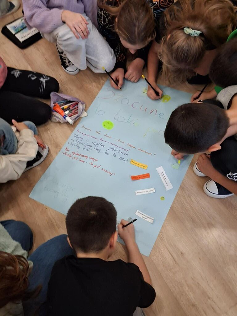 
                                                    Grupa dzieci siedzi na podłodze, pisząc i rysując na dużym niebieskim arkuszu papieru. Obok nich leżą kolorowe mazaki i kredki. Dzieci skupiają się na wspólnej pracy nad projektem związanym z szacunkiem.
                                                