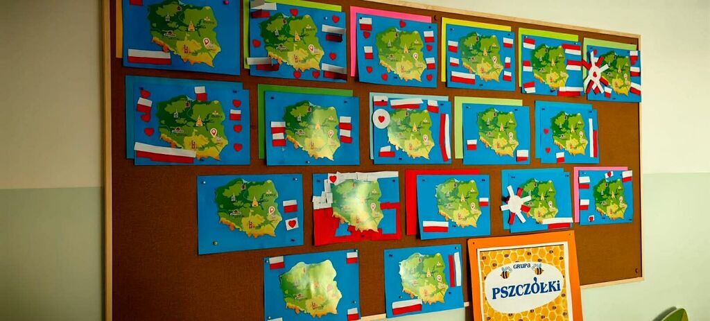 
                                                    Na tablicy ogłoszeń widoczne są kolorowe prace plastyczne przedstawiające mapy Polski z wklejonymi polskimi flagami. Pod pracami znajduje się tabliczka z napisem 
