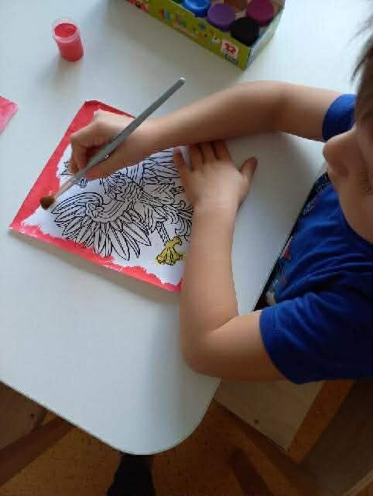 
                                                    Dziecko siedzi przy stoliku i koloruje obrazek przedstawiający ptaka. Używa pędzla i czerwonej farby do wypełnienia tła. Na stole znajdują się również przybory malarskie.
                                                