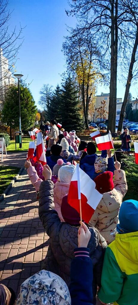 
                                                    Dzieci maszerują chodnikiem, trzymając polskie flagi. Ubrane są w ciepłe kurtki, co wskazuje na chłodną pogodę. Widok z tyłu, wokół drzewa i budynki, a nad nimi błękitne niebo.
                                                
