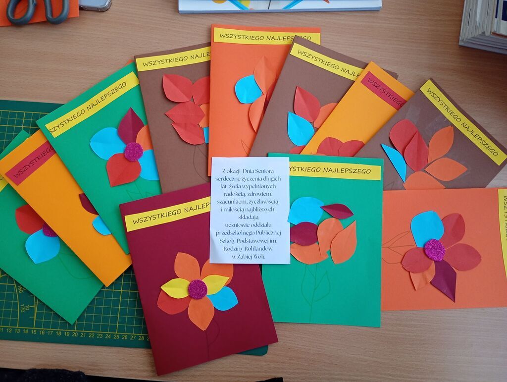 
                                                    Osiem kolorowych kartek z papieru, ozdobionych wycinankami w kształcie kwiatów. Na każdej z nich znajduje się napis „WSZYSTKIEGO NAJLEPSZEGO”. Karty są ułożone wachlarzowo na stole.
                                                