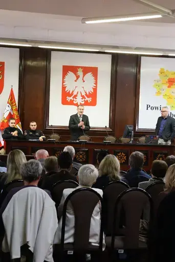 Szkolenie w sali konferencyjnej wyłożonej ciemnym drewnem: mężczyźni z mikrofonem przemawiają przy stole prezydialnym na tle godła Polski, herbu i mapy Powiatu Lubelskiego oraz flag, uczestnicy siedzą na krzesłach i słuchają, na ekranie widać slajd z napisem Zagrożenie z powietrza, szkolenie, Starostwo Powiatowe Lublin, 7 października 2026 rok, a kilkanaście osób pozuje do wspólnej fotografii na schodach podestu.