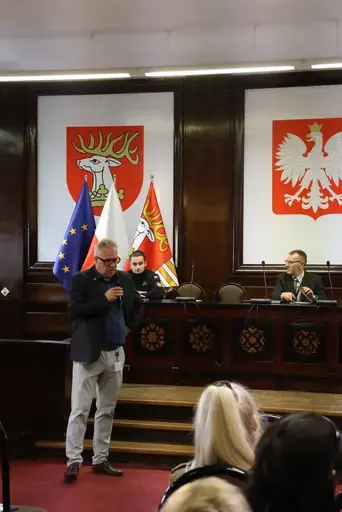 Szkolenie w sali konferencyjnej wyłożonej ciemnym drewnem: mężczyźni z mikrofonem przemawiają przy stole prezydialnym na tle godła Polski, herbu i mapy Powiatu Lubelskiego oraz flag, uczestnicy siedzą na krzesłach i słuchają, na ekranie widać slajd z napisem Zagrożenie z powietrza, szkolenie, Starostwo Powiatowe Lublin, 7 października 2026 rok, a kilkanaście osób pozuje do wspólnej fotografii na schodach podestu.