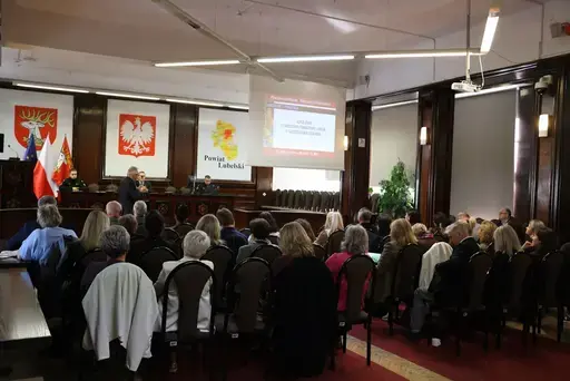 Szkolenie w sali konferencyjnej wyłożonej ciemnym drewnem: mężczyźni z mikrofonem przemawiają przy stole prezydialnym na tle godła Polski, herbu i mapy Powiatu Lubelskiego oraz flag, uczestnicy siedzą na krzesłach i słuchają, na ekranie widać slajd z napisem Zagrożenie z powietrza, szkolenie, Starostwo Powiatowe Lublin, 7 października 2026 rok, a kilkanaście osób pozuje do wspólnej fotografii na schodach podestu.