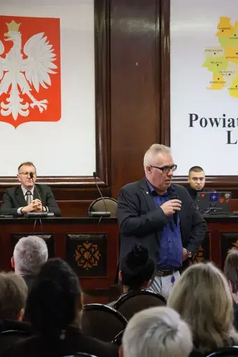 Szkolenie w sali konferencyjnej wyłożonej ciemnym drewnem: mężczyźni z mikrofonem przemawiają przy stole prezydialnym na tle godła Polski, herbu i mapy Powiatu Lubelskiego oraz flag, uczestnicy siedzą na krzesłach i słuchają, na ekranie widać slajd z napisem Zagrożenie z powietrza, szkolenie, Starostwo Powiatowe Lublin, 7 października 2026 rok, a kilkanaście osób pozuje do wspólnej fotografii na schodach podestu.