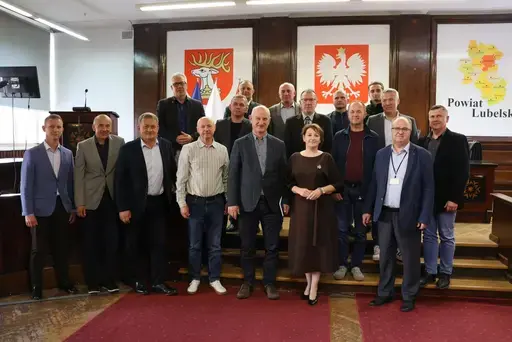 Szkolenie w sali konferencyjnej wyłożonej ciemnym drewnem: mężczyźni z mikrofonem przemawiają przy stole prezydialnym na tle godła Polski, herbu i mapy Powiatu Lubelskiego oraz flag, uczestnicy siedzą na krzesłach i słuchają, na ekranie widać slajd z napisem Zagrożenie z powietrza, szkolenie, Starostwo Powiatowe Lublin, 7 października 2026 rok, a kilkanaście osób pozuje do wspólnej fotografii na schodach podestu.