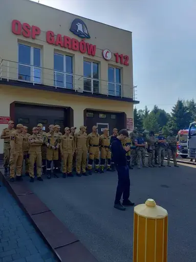 Ćwiczenia służb na placu przed remizą z napisem OSP Garbów 112: strażacy w piaskowych mundurach stoją w szeregu, obok funkcjonariusze w kamuflażu i kominiarkach, trzy osoby w żółtych kombinezonach ochronnych z maskami i zielonymi rękawicami, czerwony wóz strażacki obok rozstawionego pomarańczowego namiotu pneumatycznego, w tle pojazdy służb i wysokie świerki pod bezchmurnym niebem.