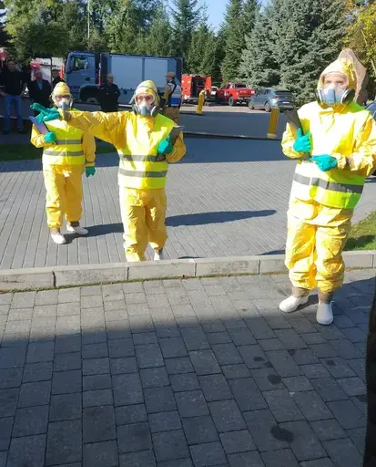 Ćwiczenia służb na placu przed remizą z napisem OSP Garbów 112: strażacy w piaskowych mundurach stoją w szeregu, obok funkcjonariusze w kamuflażu i kominiarkach, trzy osoby w żółtych kombinezonach ochronnych z maskami i zielonymi rękawicami, czerwony wóz strażacki obok rozstawionego pomarańczowego namiotu pneumatycznego, w tle pojazdy służb i wysokie świerki pod bezchmurnym niebem.