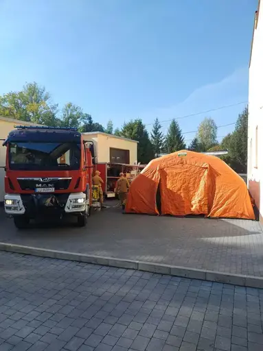 Ćwiczenia służb na placu przed remizą z napisem OSP Garbów 112: strażacy w piaskowych mundurach stoją w szeregu, obok funkcjonariusze w kamuflażu i kominiarkach, trzy osoby w żółtych kombinezonach ochronnych z maskami i zielonymi rękawicami, czerwony wóz strażacki obok rozstawionego pomarańczowego namiotu pneumatycznego, w tle pojazdy służb i wysokie świerki pod bezchmurnym niebem.