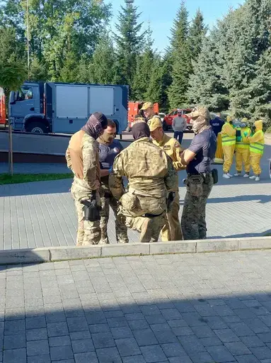 Ćwiczenia służb na placu przed remizą z napisem OSP Garbów 112: strażacy w piaskowych mundurach stoją w szeregu, obok funkcjonariusze w kamuflażu i kominiarkach, trzy osoby w żółtych kombinezonach ochronnych z maskami i zielonymi rękawicami, czerwony wóz strażacki obok rozstawionego pomarańczowego namiotu pneumatycznego, w tle pojazdy służb i wysokie świerki pod bezchmurnym niebem.