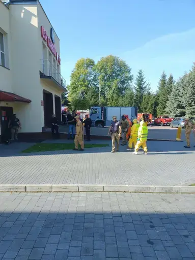 Ćwiczenia służb na placu przed remizą z napisem OSP Garbów 112: strażacy w piaskowych mundurach stoją w szeregu, obok funkcjonariusze w kamuflażu i kominiarkach, trzy osoby w żółtych kombinezonach ochronnych z maskami i zielonymi rękawicami, czerwony wóz strażacki obok rozstawionego pomarańczowego namiotu pneumatycznego, w tle pojazdy służb i wysokie świerki pod bezchmurnym niebem.