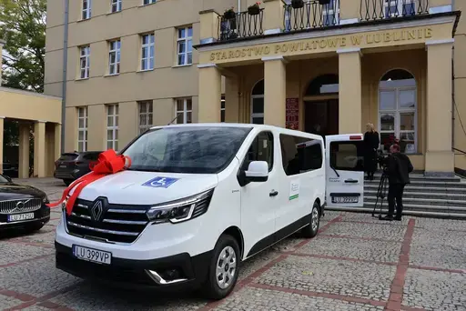 Przekazanie białego busa Renault Trafic z czerwoną kokardą przed budynkiem Starostwa Powiatowego w Lublinie: winda w tylnych drzwiach, wywiady dla telewizji, wspólne zdjęcia z podopiecznymi i spotkanie przy stole w sali.