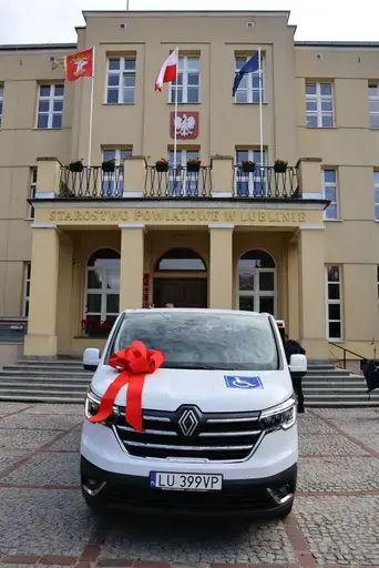Przekazanie białego busa Renault Trafic z czerwoną kokardą przed budynkiem Starostwa Powiatowego w Lublinie: winda w tylnych drzwiach, wywiady dla telewizji, wspólne zdjęcia z podopiecznymi i spotkanie przy stole w sali.