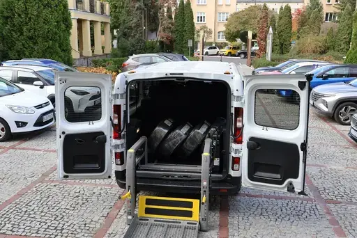 Przekazanie białego busa Renault Trafic z czerwoną kokardą przed budynkiem Starostwa Powiatowego w Lublinie: winda w tylnych drzwiach, wywiady dla telewizji, wspólne zdjęcia z podopiecznymi i spotkanie przy stole w sali.