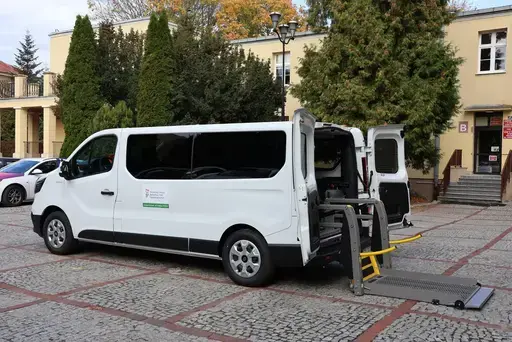 Przekazanie białego busa Renault Trafic z czerwoną kokardą przed budynkiem Starostwa Powiatowego w Lublinie: winda w tylnych drzwiach, wywiady dla telewizji, wspólne zdjęcia z podopiecznymi i spotkanie przy stole w sali.