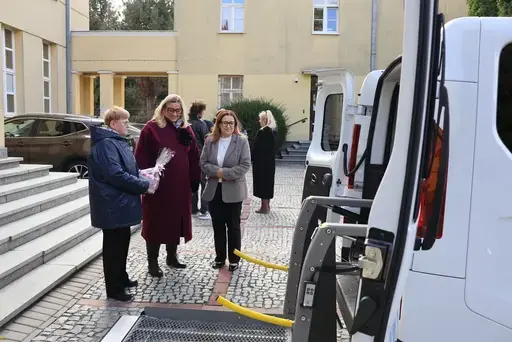Przekazanie białego busa Renault Trafic z czerwoną kokardą przed budynkiem Starostwa Powiatowego w Lublinie: winda w tylnych drzwiach, wywiady dla telewizji, wspólne zdjęcia z podopiecznymi i spotkanie przy stole w sali.