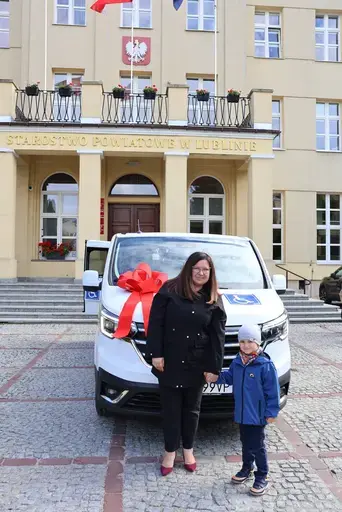 Przekazanie białego busa Renault Trafic z czerwoną kokardą przed budynkiem Starostwa Powiatowego w Lublinie: winda w tylnych drzwiach, wywiady dla telewizji, wspólne zdjęcia z podopiecznymi i spotkanie przy stole w sali.