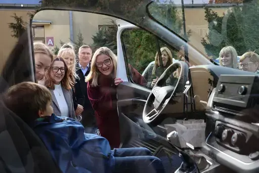 Przekazanie białego busa Renault Trafic z czerwoną kokardą przed budynkiem Starostwa Powiatowego w Lublinie: winda w tylnych drzwiach, wywiady dla telewizji, wspólne zdjęcia z podopiecznymi i spotkanie przy stole w sali.