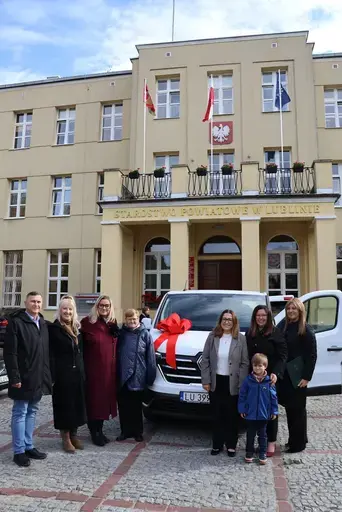 Przekazanie białego busa Renault Trafic z czerwoną kokardą przed budynkiem Starostwa Powiatowego w Lublinie: winda w tylnych drzwiach, wywiady dla telewizji, wspólne zdjęcia z podopiecznymi i spotkanie przy stole w sali.