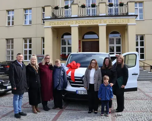 Przekazanie białego busa Renault Trafic z czerwoną kokardą przed budynkiem Starostwa Powiatowego w Lublinie: winda w tylnych drzwiach, wywiady dla telewizji, wspólne zdjęcia z podopiecznymi i spotkanie przy stole w sali.