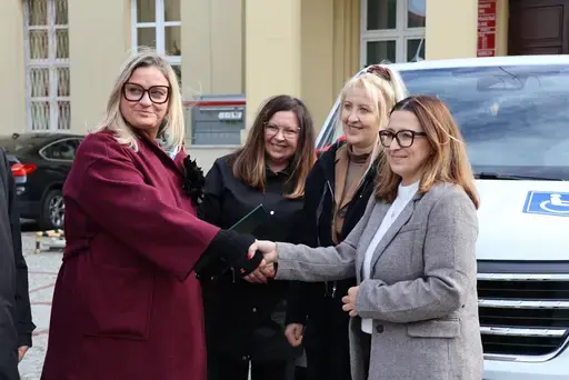 Przekazanie białego busa Renault Trafic z czerwoną kokardą przed budynkiem Starostwa Powiatowego w Lublinie: winda w tylnych drzwiach, wywiady dla telewizji, wspólne zdjęcia z podopiecznymi i spotkanie przy stole w sali.
