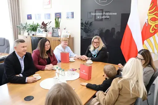 Przekazanie białego busa Renault Trafic z czerwoną kokardą przed budynkiem Starostwa Powiatowego w Lublinie: winda w tylnych drzwiach, wywiady dla telewizji, wspólne zdjęcia z podopiecznymi i spotkanie przy stole w sali.