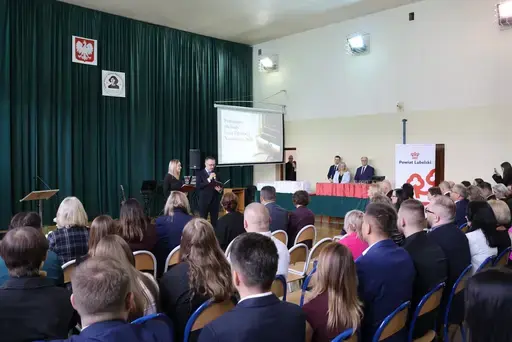 Obchody Powiatowego Dnia Edukacji Narodowej w sali gimnastycznej szkoły w Bełżycach: wystąpienia przy mównicy, wręczanie dyplomów i upominków nauczycielom oraz uczniom, goście na widowni, poczet sztandarowy i występ muzyczny uczniów.