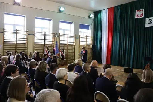 Obchody Powiatowego Dnia Edukacji Narodowej w sali gimnastycznej szkoły w Bełżycach: wystąpienia przy mównicy, wręczanie dyplomów i upominków nauczycielom oraz uczniom, goście na widowni, poczet sztandarowy i występ muzyczny uczniów.